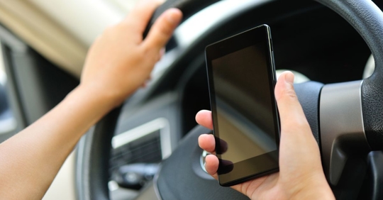 Man holding a cell phone in right hand while steering a car with his left hand.
