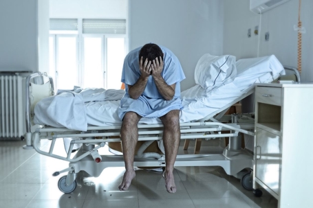 Man in hospital sitting on side of bed, holding head in hands